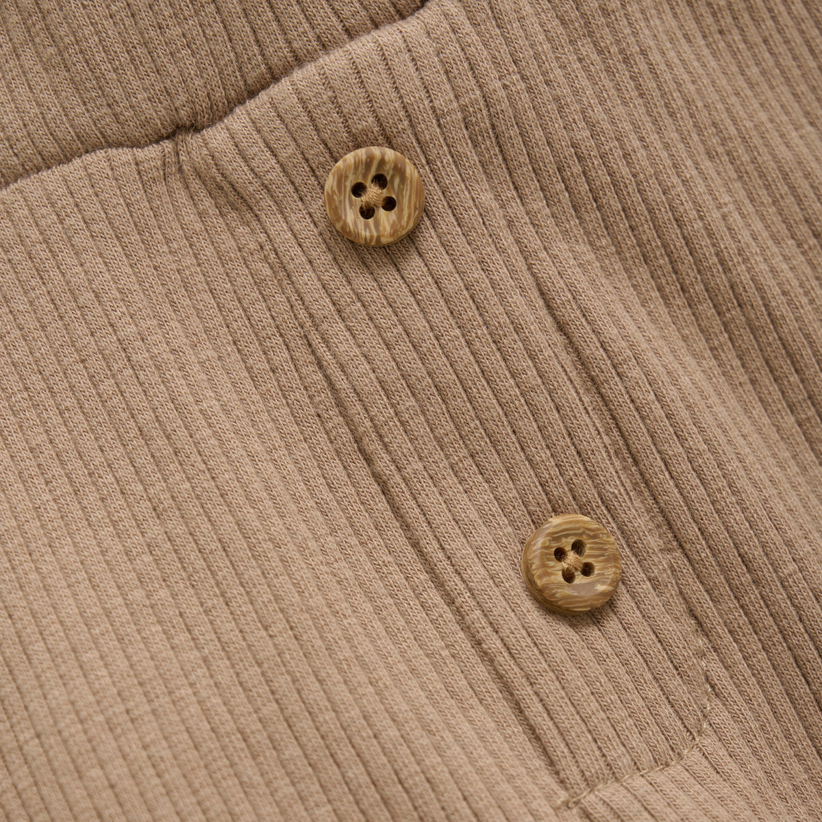 Nahaufnahme eines beigen Rippstoffs mit zwei braunen Knöpfen mit jeweils vier Löchern, die auf den Stoff genäht sind und seine Struktur und Details hervorheben. Der weiche Stoff aus TENCEL™ Modal-Fasern passt perfekt zu den gemütlichen Minymo - Kurze Hosen aus Ripp-Stoff mit einem elastischen Bund für zusätzlichen Komfort von Minymo.