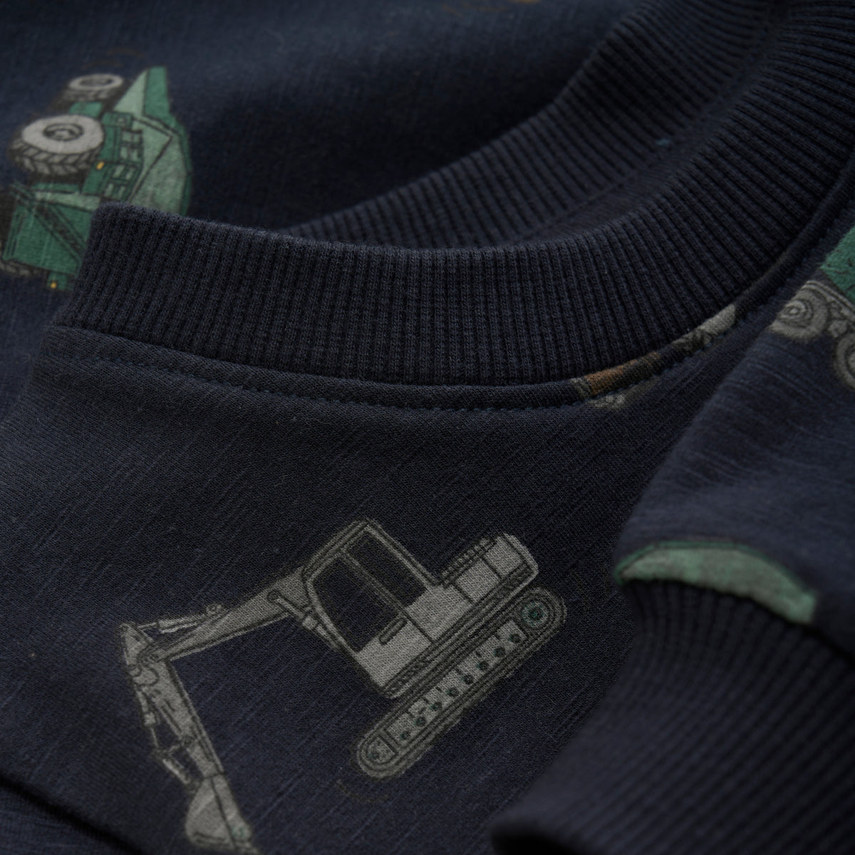 Nahaufnahme eines dunkelblauen Stoffes mit einem Baufahrzeug-Druck, der Bagger und Muldenkipper zeigt. Der gerippte Kragen und Ärmelsaum dieses Minymo-Pullovers von Minymo sind sichtbar, gefertigt aus einer weichen Baumwollmischung mit subtiler Textur.