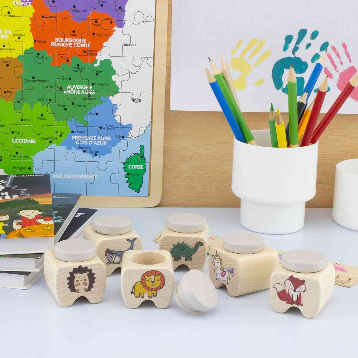 Ein farbenfroher Kinderschreibtisch mit einer Puzzlekarte von Frankreich an der Wand, einem weißen Becher voller Buntstifte, Handabdruckkunst und mehreren kleinen Holzbehältern mit verschiedenen Tiermotiven wie Igel, Löwe und Fuchs. Verstreute Hefte ergänzen die Zahnbox Tiere von Ulysse Couleurs d'Enfance, um die Erinnerungen Ihres Kindes an den Verlust der Milchzähne zu bewahren.