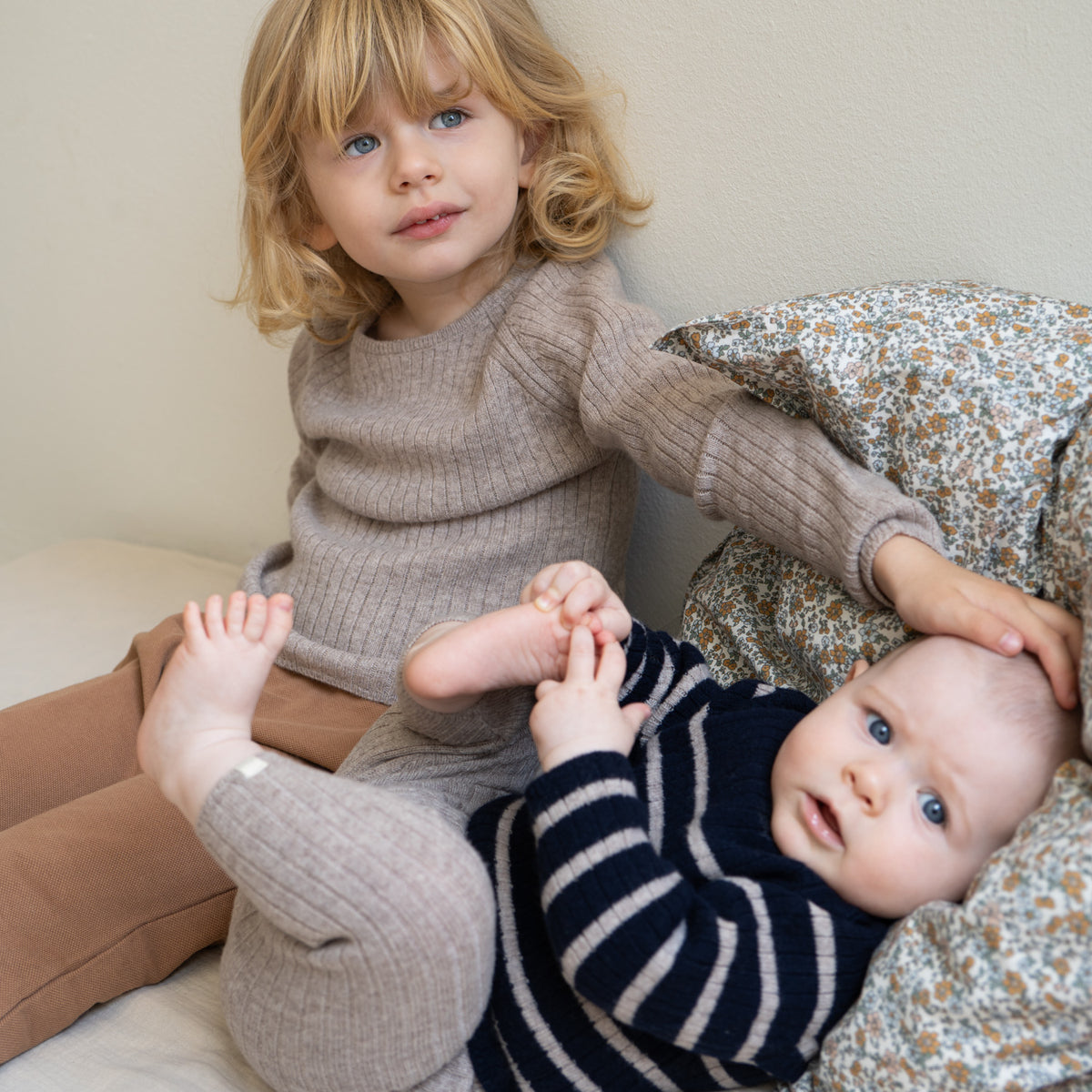 Zwei kleine Kinder sitzen auf einem Bett. Das ältere Kind trägt einen beigefarbenen Strickpullover und Hosen aus Materialien nach dem Responsible Wool Standard und blickt mit einem leichten Lächeln in die Kamera. Das Baby liegt neben dem älteren Kind in Minimalismas Langarm-Shirt (Kopie), einer marineblau gestreiften Thermoschicht, und blickt ebenfalls mit einem neugierigen Gesichtsausdruck in die Kamera.
