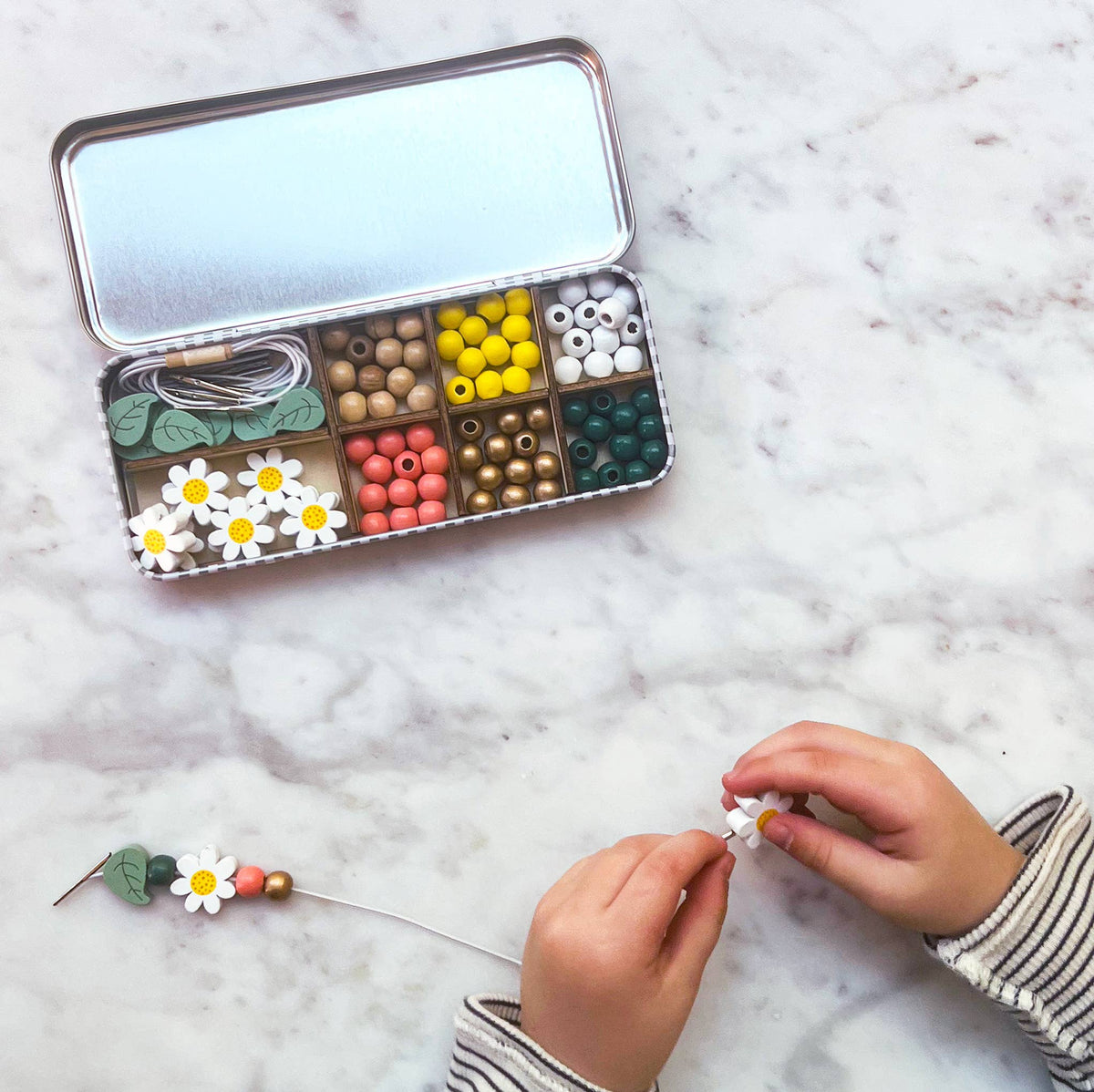 Man sieht, wie Kinderhände eine Perle auf eine Schnur fädeln und so ein Armband mit Gänseblümchenmotiv herstellen. Die Aktivität, die durch das Cotton Twist - DIY Perlen-Set Armband von Cotton Twist erleichtert wird, verbessert die Feinmotorik und wird neben einer offenen Blechdose auf einer Marmoroberfläche durchgeführt, die verschiedene plastikfreie Bastelperlen, Schnüre und Gänseblümchendekorationen enthält, die ordentlich in Fächern angeordnet sind.