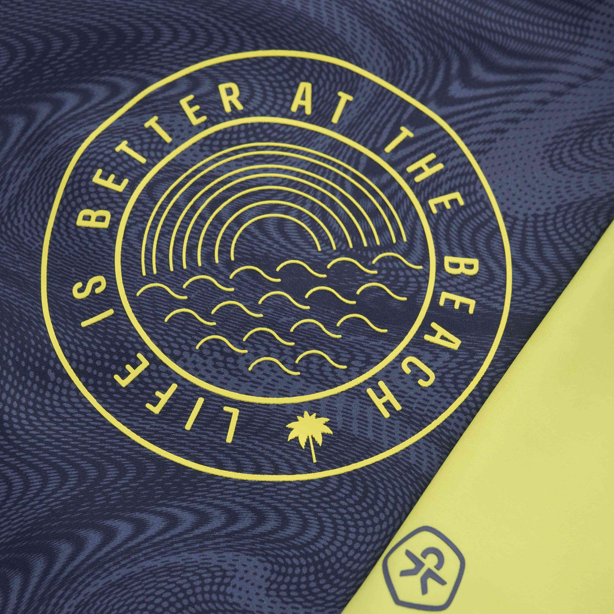 Eine Nahaufnahme eines blauen Stoffes mit einer kreisförmigen gelben Grafik mit den Worten „Life is Better at the Beach“. Innerhalb des Kreises befindet sich eine abstrakte Darstellung von Wellen, einer Sonne und einer kleinen Palme. Auf diesem Color Kids - UV Shirt langarm von Color Kids ist auch ein kleineres Logo einer laufenden Strichmännchenfigur zu sehen.