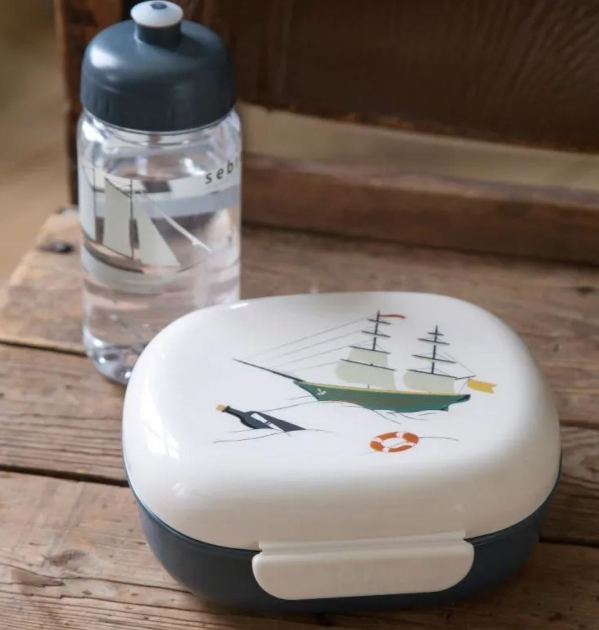 Eine Kinderwasserflasche und eine Sebra-Lunchbox mit nautischem Motiv stehen auf einer Holzoberfläche. Die Kinderflaschen-Thermoflasche ist mit einem Segelbootmotiv versehen, während die Lunchbox ein Schiff, einen Anker und einen Rettungsring zeigt.