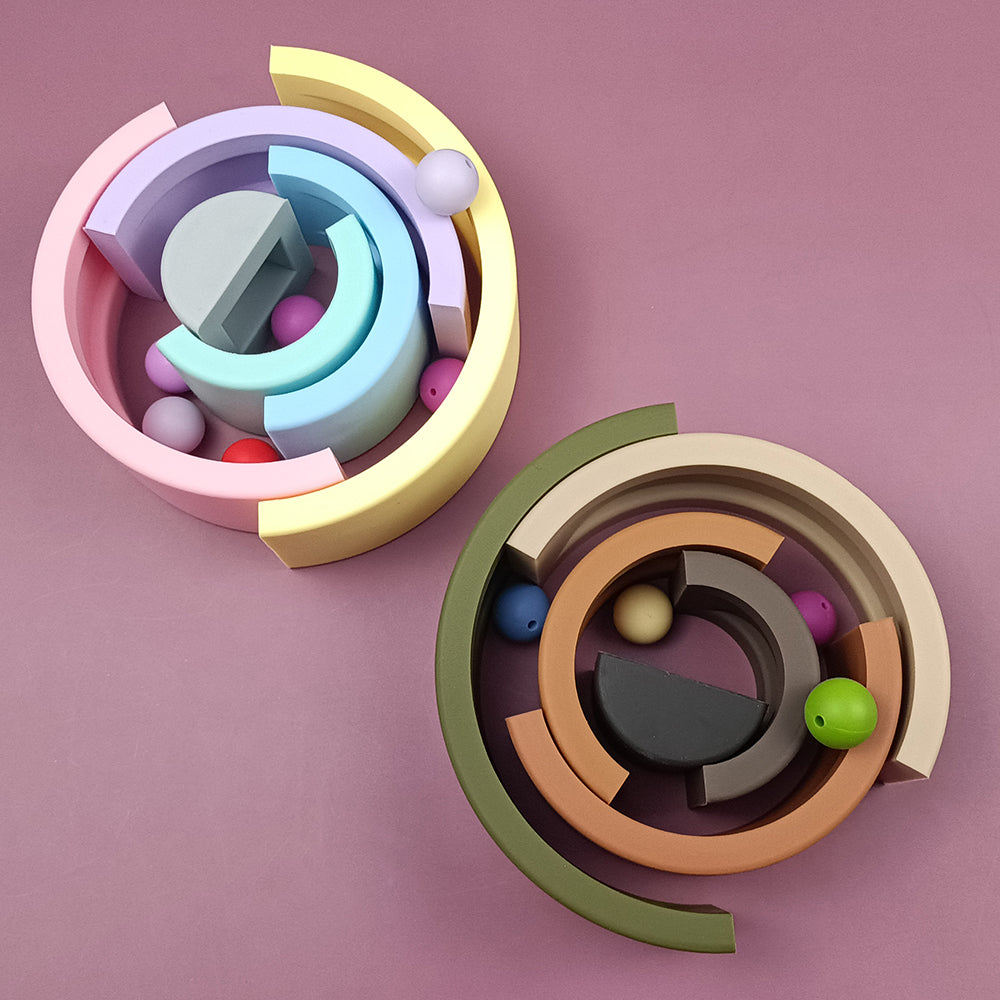 Das Bild zeigt Eric Baby - Steckspielzeug Regenbogen 6-teilig aus Silikon, zwei runde, mehrfarbige Holzpuzzlespielzeuge auf rosa Hintergrund. Jedes Spielzeug besteht aus ineinandergreifenden, gebogenen Teilen und kleinen, bunten Bällen, die zwischen den Segmenten eingebettet sind. Daneben befindet sich eine glänzende Kindergarten-Brotdose aus Edelstahl, die der spielerischen Szene eine moderne Note verleiht.