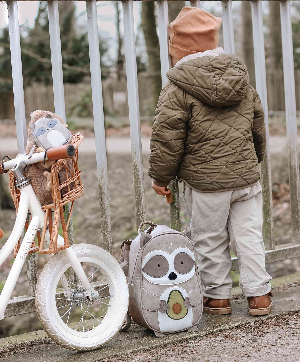 Ein Kind in einer braunen Steppjacke und hellbraunen Hosen schaut durch einen Zaun. Neben dem Kind steht ein weißes Laufrad mit einem Plüschtier im Korb, geschmückt mit spülmaschinenfesten Aufklebern. Auf dem Boden liegt ein Little Who - Rucksack Waschbär Waldemar von Little Who, mit einem Cartoon-Eulen-Design. Die Szene spielt im Freien mit Bäumen im Hintergrund.