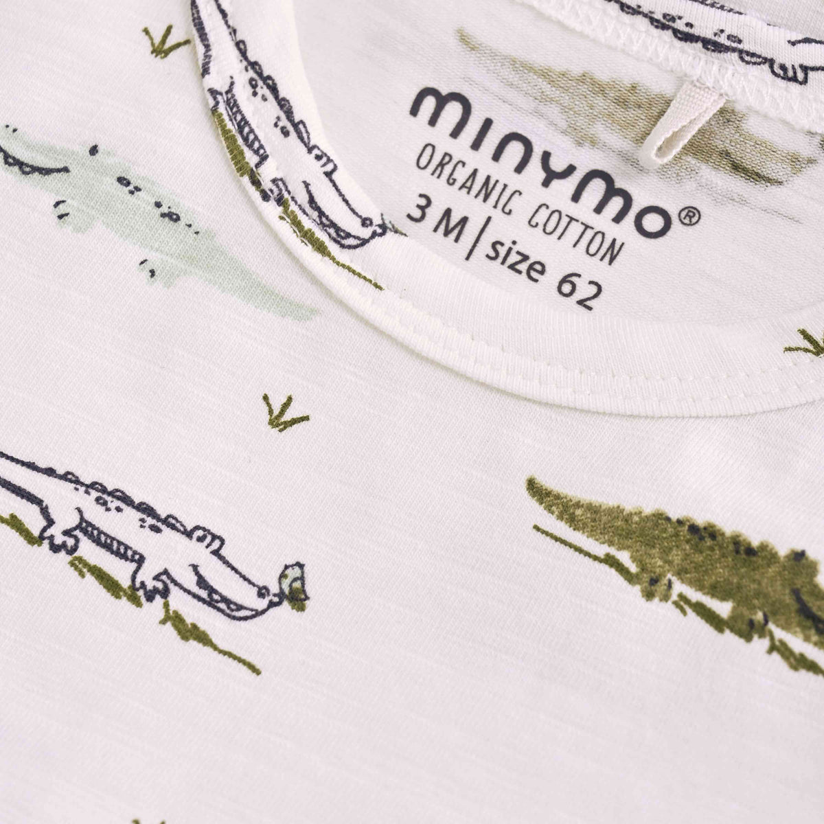 Nahaufnahme eines Babybodys aus weißem Stoff mit grünem Krokodilmuster. Auf dem Etikett steht in schwarzer Schrift „Minymo - Body kurzarm mit Allover-Print 3M Größe 62“. Der Stoff wirkt weich und gut verarbeitet, passend für ein Baby.