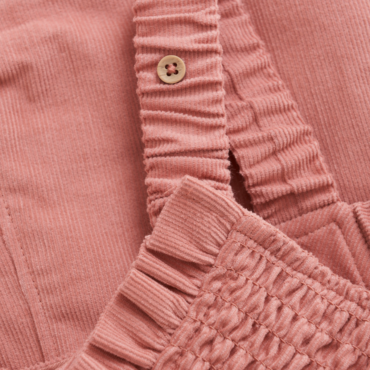 Nahaufnahme des Minymo - Overalls, einem rosa Cord-Kleidungsstück mit einem Bund mit gerafften elastischen Details und einem einzelnen hellbraunen Knopf. Der weiche und gemütliche Stoff besteht aus 100 % Baumwolle und hat eine gerippte Textur. Dieses Stück ist GOTS-zertifiziert, was seine Nachhaltigkeit und Qualität gewährleistet.