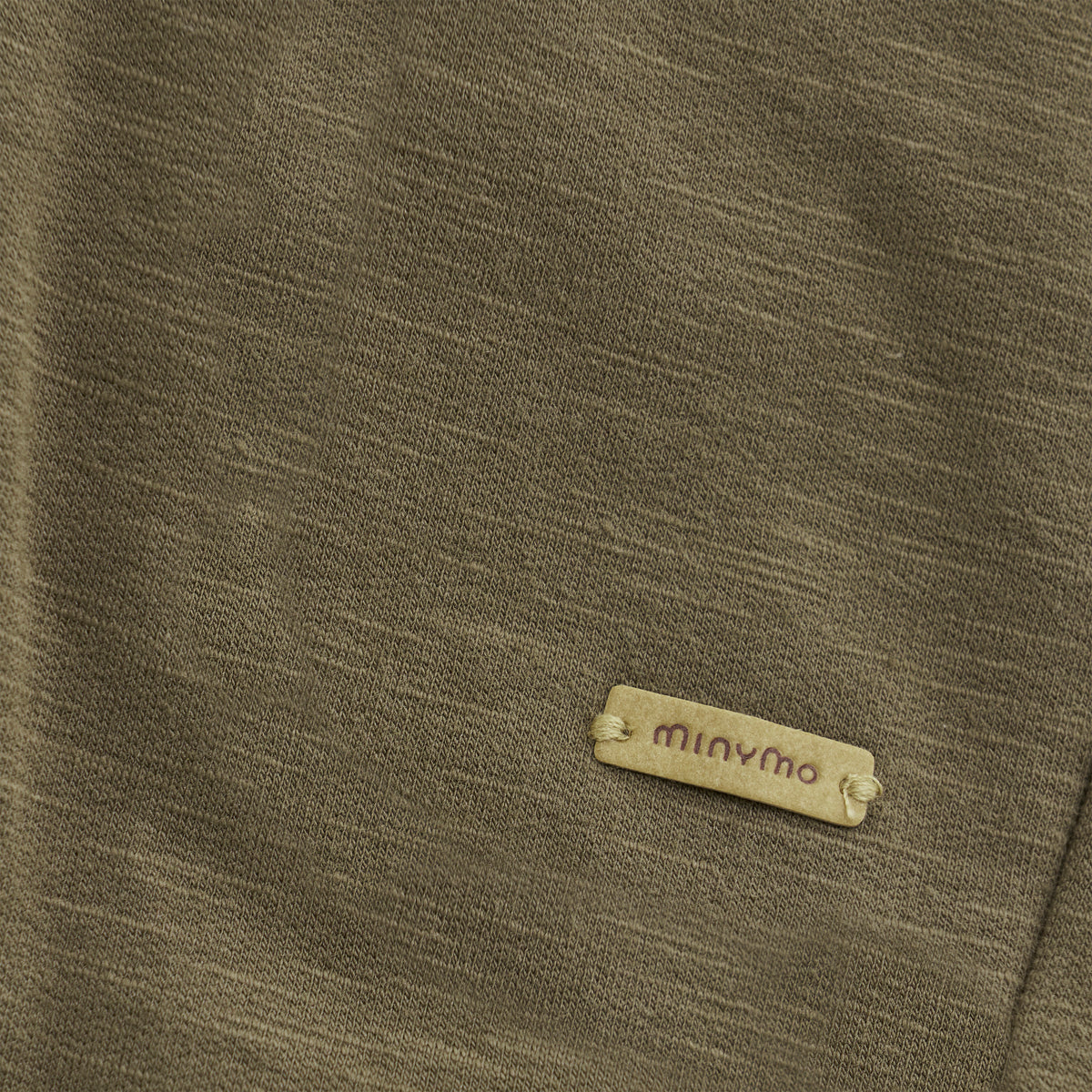 Eine Nahaufnahme eines khakifarbenen Stoffes, wahrscheinlich von einer Minymo-Hose, mit einem kleinen beigefarbenen Etikett mit der Aufschrift „minymo“. Der Stoff aus weicher Baumwollmischung wirkt weich und leicht strukturiert. Das rechteckige Etikett mit abgerundeten Kanten zeigt den Markennamen in Kleinbuchstaben.