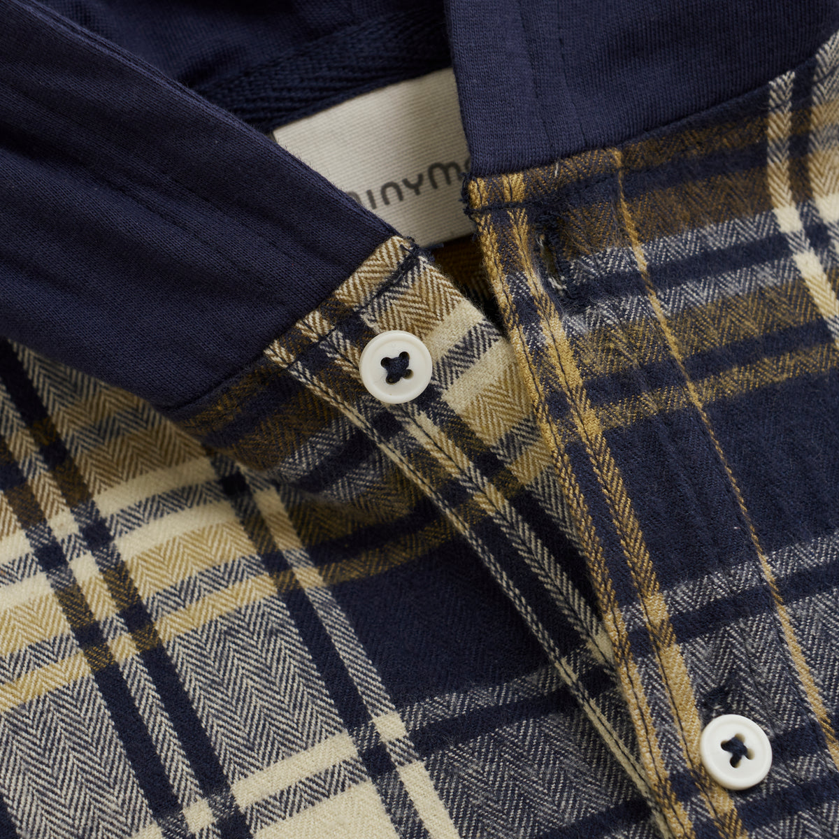 Nahaufnahme des Minymo - Hoodie Hemdes mit einem karierten Muster in Marineblau, Senfgelb und Creme. Das Hemd besteht aus 100 % Baumwolle und hat einen Button-Down-Kragen mit weißen Knöpfen. Im Kragenbereich ist ein Teiletikett sichtbar, das das Minymo-Branding hervorhebt.