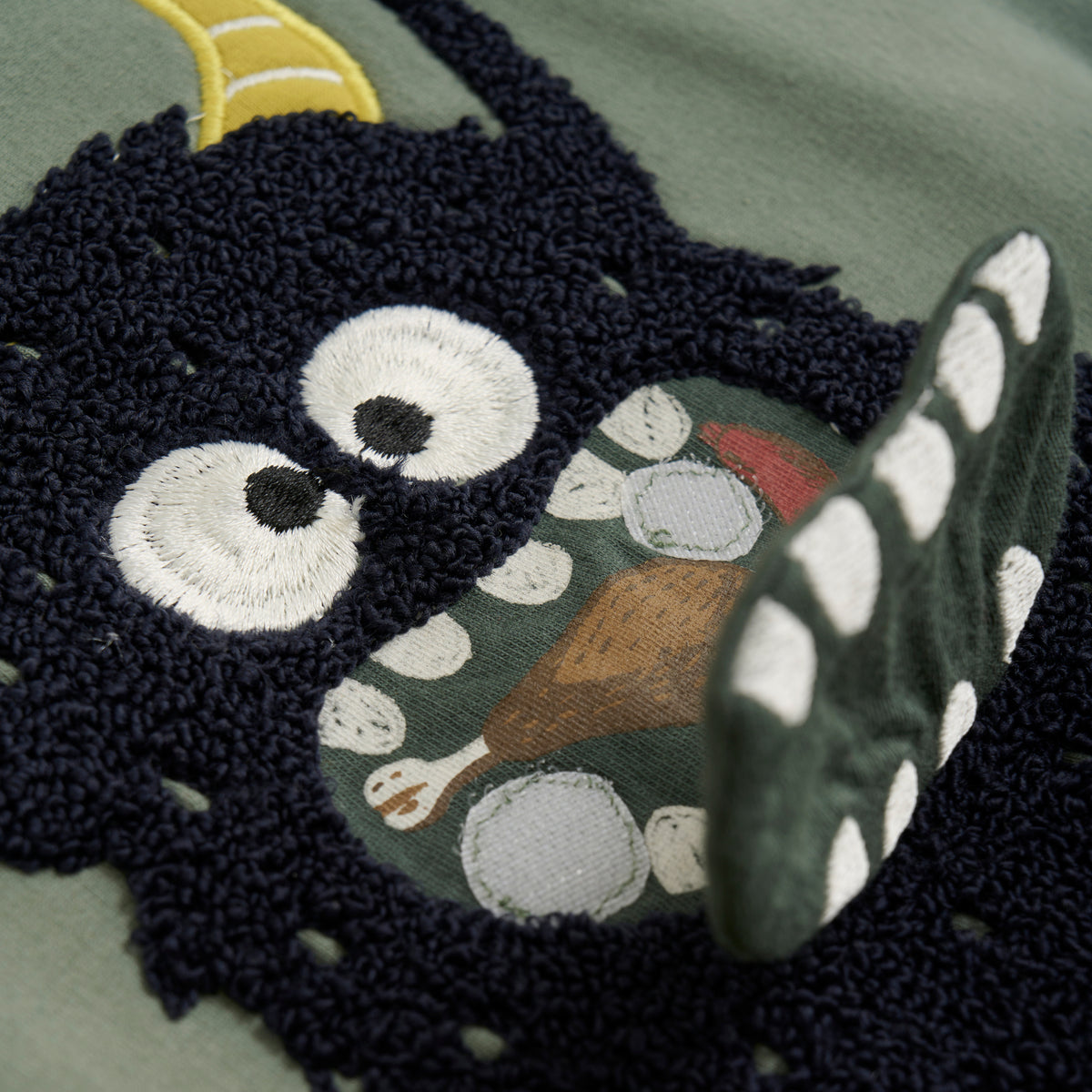 Nahaufnahme eines Stoffdesigns auf einem GOTS-zertifizierten Minymo-Pullover, der eine skurrile dunkelblaue Kreatur mit großen gestickten Augen und offenem Mund zeigt. Im Mund ist ein Bild von Nahrungsmitteln, darunter eine Hähnchenkeule, vor einem grünen Hintergrund zu sehen. Die Kreatur hat gelbe Akzente, die an Hörner erinnern.
