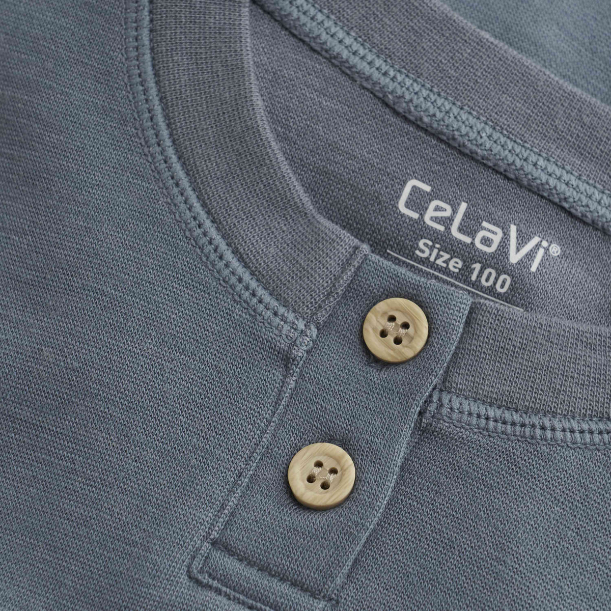 Nahaufnahme einer blaugrauen CeLaVi Bluse mit Rundhalsausschnitt, zwei beigen Knöpfen unter dem Kragen und einem sichtbaren Etikett mit der Marke CeLaVi und der Größe 100. Der Stoff hat eine weiche, strukturierte Optik und erfüllt die Qualitätskriterien des OEKO-TEX STANDARD 100.