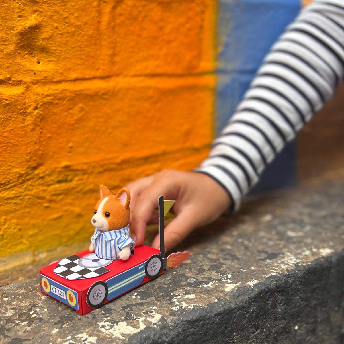 Eine Hand mit einem gestreiften Langarmshirt schiebt den Cotton Twist - DIY Matchbox-Rennwagen, auf dem eine Figur zu sehen ist, die einem Hund in passendem Outfit ähnelt. Die Szene spielt vor dem bezaubernden Hintergrund einer bunt bemalten Backsteinmauer mit orangefarbenen und blauen Abschnitten und fängt perfekt den spielerischen Geist ein, der mühelos Feinmotorik fördert.