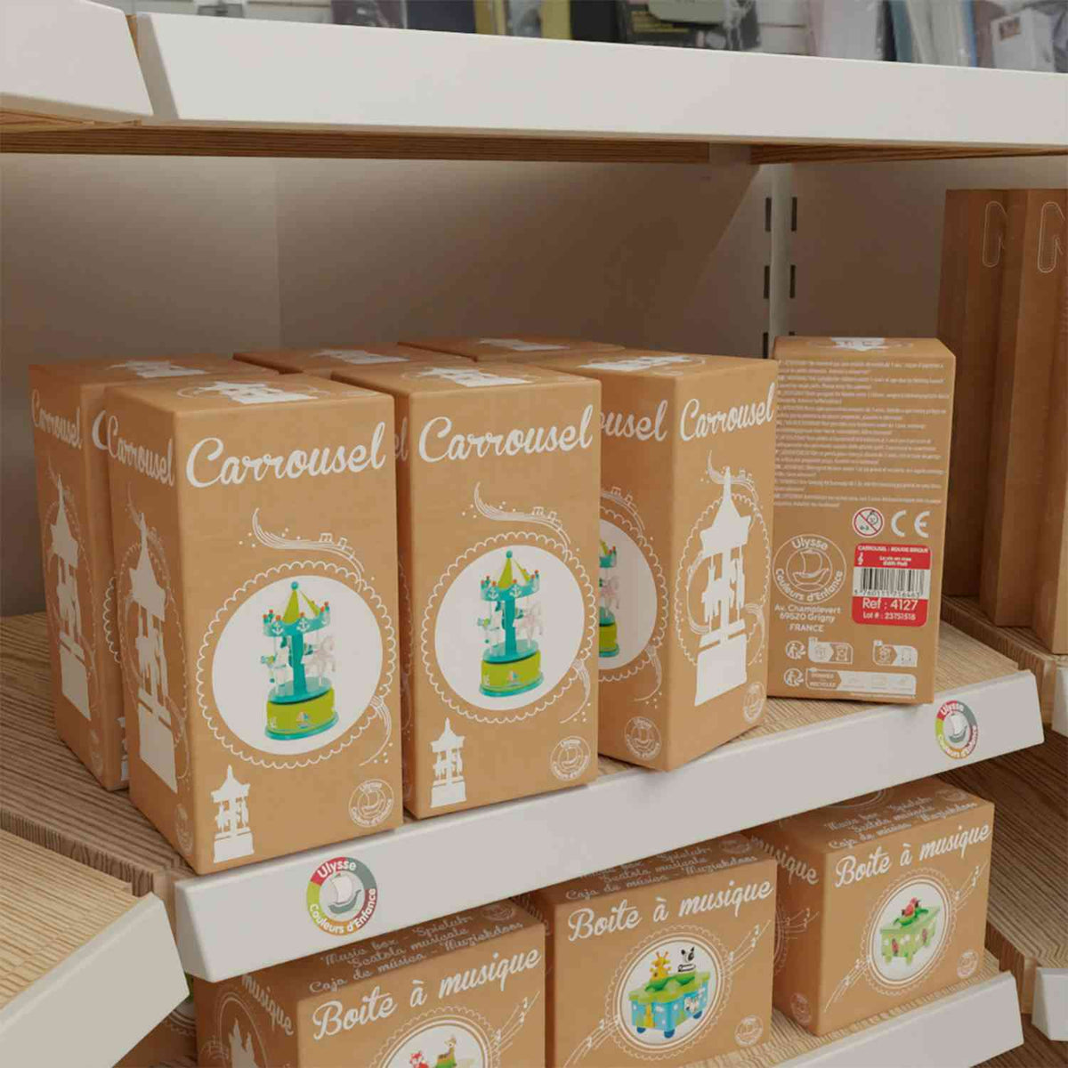 Ein Ladenregal mit mehreren verpackten Ulysse Couleurs d'Enfance - Karussells in Grün mit einem grün-gelben Design. Die braune Verpackung zeigt eine Abbildung des Karussells und den Text „Carrousel“ auf der Vorderseite. Auf der Seite einer der Schachteln ist ein Etikett mit Anweisungen zu sehen, perfekt als pädagogisches Spielzeug ab 2 von Ulysse Couleurs d'Enfance.