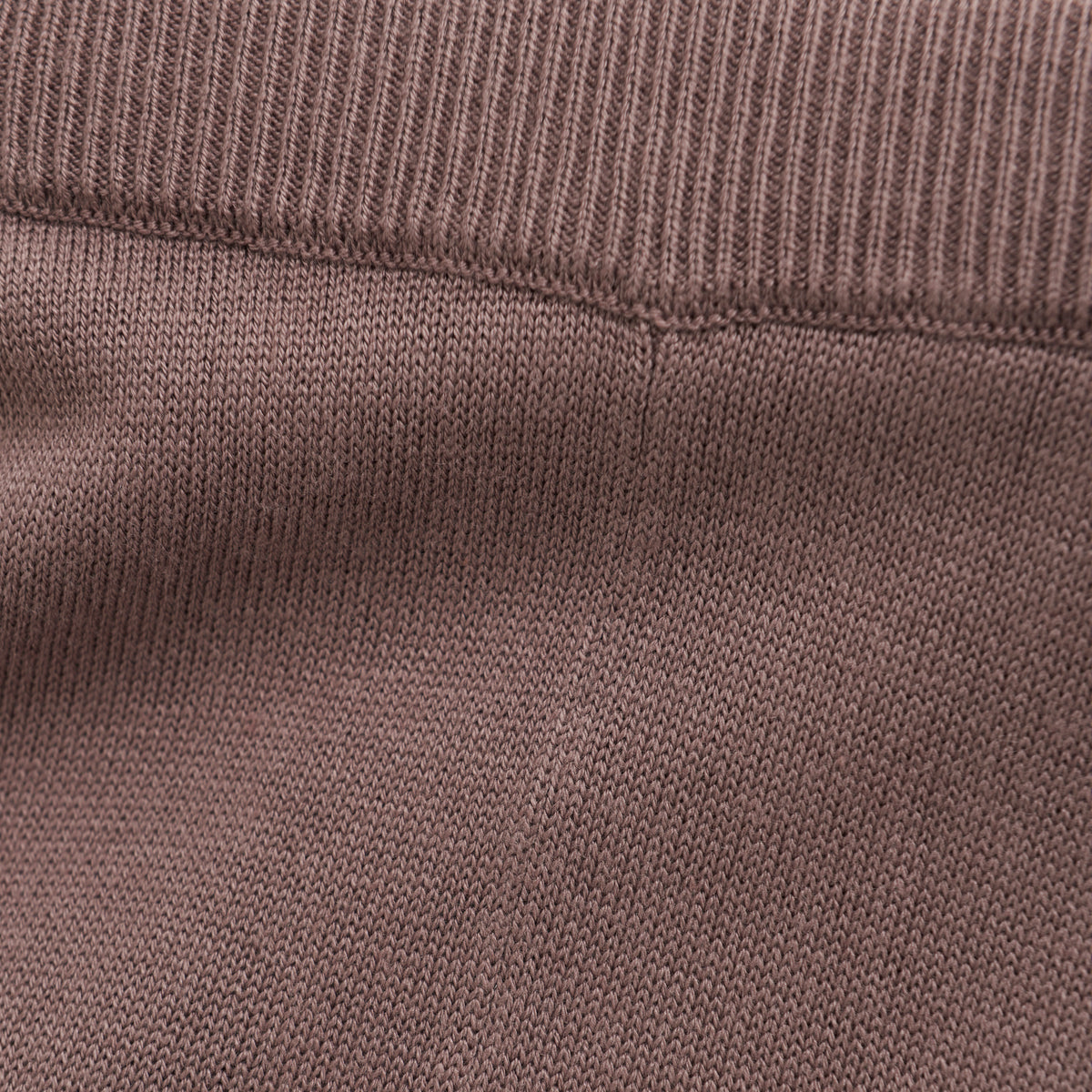 Nahaufnahme eines Abschnitts braunen Strickstoffs. Das Bild zeigt eine Naht, an der zwei Teile zusammengefügt sind, wodurch die Textur und die Nähte hervorgehoben werden. Das Oberteil hat ein geripptes Muster, das an den Gummizug in der Taille erinnert, der bei Fixoni – Hosen von Fixoni zu sehen ist, während das Unterteil ein glatteres Strickmuster aufweist.