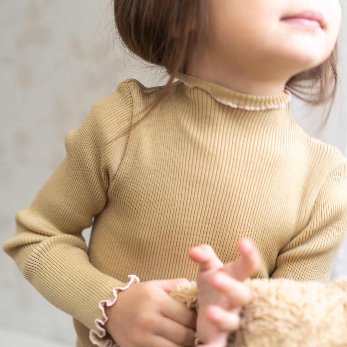 Ein kleines Kind trägt ein geripptes, langärmeliges Langarmshirt aus beigefarbener Merinowolle mit Rüschenmanschetten und -kragen und hält einen Plüschteddybär. Das Gesicht des Kindes ist teilweise sichtbar und blickt zur linken Seite des Bildes.