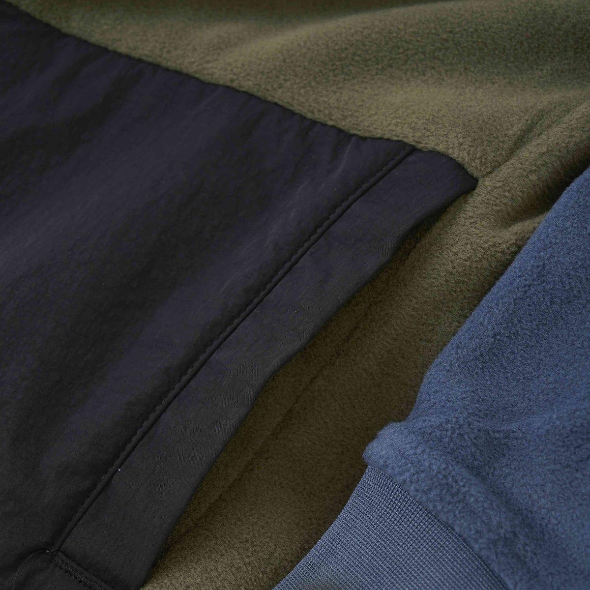 Nahaufnahme der Color Kids - Fleecejacke - Colorblock von Color Kids, die drei verschiedene Stoffe und Farben zeigt. Der linke Abschnitt besteht aus olivgrünem Fleece, der mittlere Abschnitt aus schwarzem Synthetikmaterial mit sichtbaren Nähten und der rechte Abschnitt aus blauem Fleece. Die Texturen sind glatt und gemütlich.