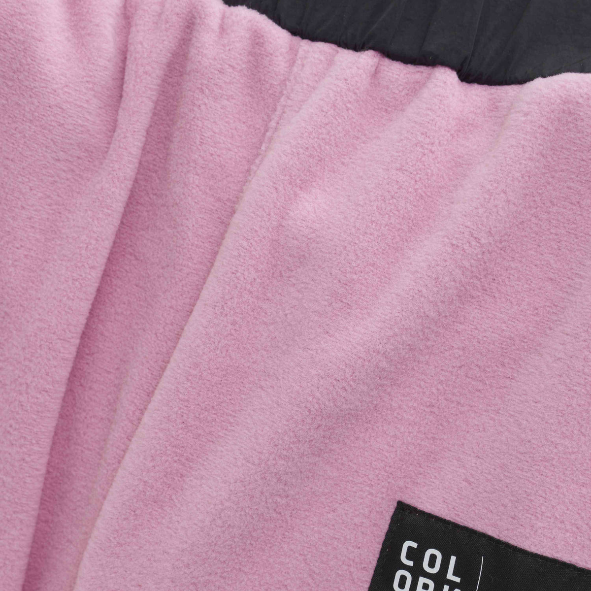 Nahaufnahme eines weichen, rosa Stoffs „Color Kids - Fleece Hosen“, auf den ein Teil eines schwarzen Etiketts aufgenäht ist. Auf dem Etikett ist teilweise weisser Text zu sehen, der die Buchstaben „COL OR“ zeigt. Am oberen Bildrand ist die Oberkante eines dunkleren Stoffs zu sehen.