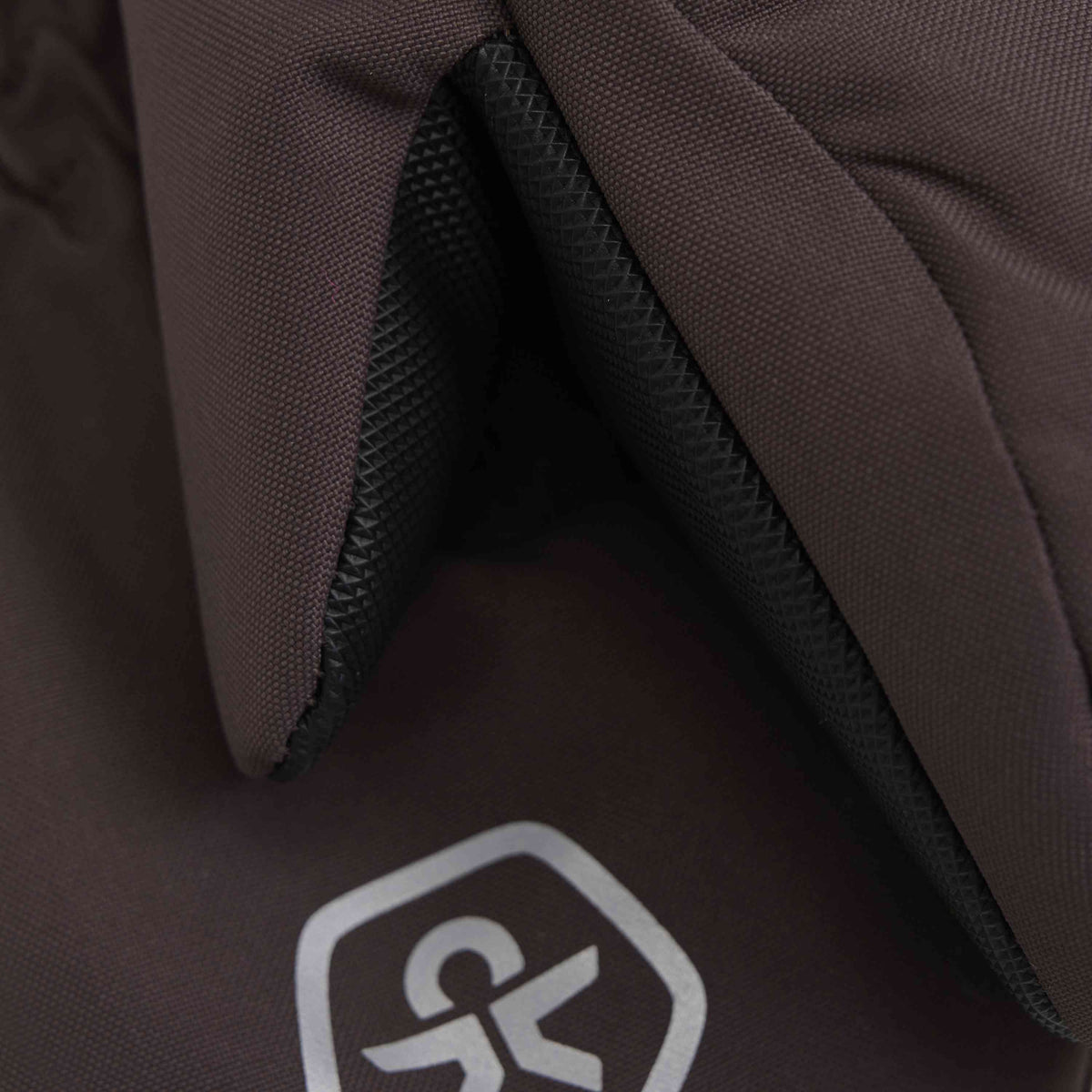 Nahaufnahme eines Paars brauner Stoffhandschuhe, wobei der Fokus auf der Naht liegt, wo der Reissverschluss auf das Material trifft. Der Stoff besteht aus recycelten Materialien, hat eine strukturierte Optik und ist wind- und wasserdicht. Unten in der Mitte befindet sich ein kleines, silbernes abstraktes Logo.