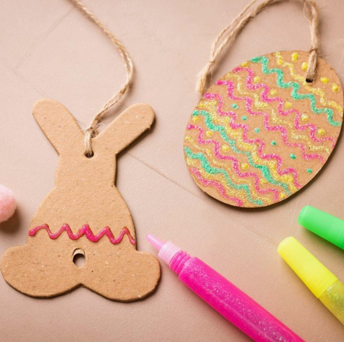 Es werden zwei Bastelarbeiten aus Karton mit Ostermotiven ausgestellt, die sich perfekt für eine Creative Company eignen – Oster-DIY-Set für Kinder – Ei und Hase. Eines hat die Form eines Häschens mit rosa Glitzer und einer daran befestigten Schnur. Das andere ist ein buntes Ei mit Wellenmuster und grünem, rosa und gelbem Glitzer. Neonpinke und grüne Glitzerstifte liegen in der Nähe – ideales Bastelbedarf für Kinder von Creative Company.