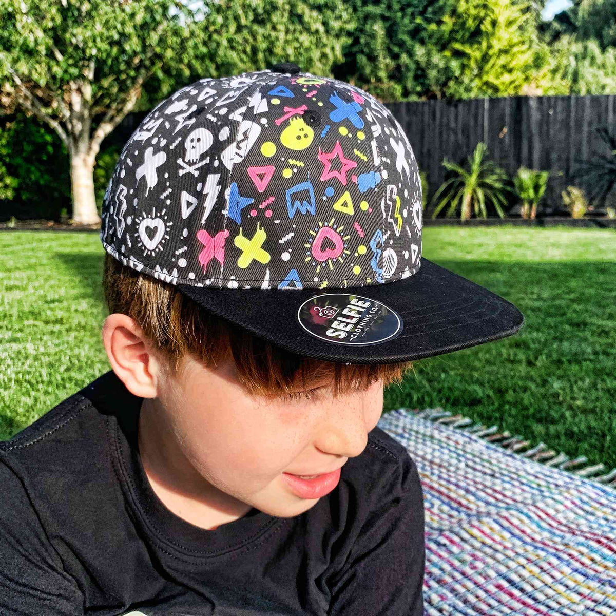 Ein Kind mit braunen Haaren sitzt auf einer bunten Decke im Gras und ist in sein Bastelset für Kinder vertieft. Es trägt eine Selfie Craft Co. - Graffiti Colour im Snapback Kappe von Selfie Craft mit bunten Cartoon-Symbolen wie Totenköpfen, Blitzen, Herzen und Sternen. Der Hintergrund zeigt einen grünen Rasen mit Bäumen und Pflanzen.