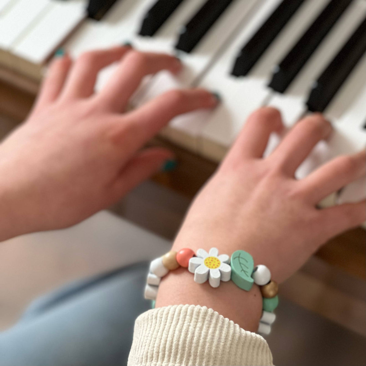 Eine Person trägt ein farbenfrohes Armband aus dem Cotton Twist - DIY Armbandset Gänseblümchen von Cotton Twist und spielt Klavier. Ihre Hände liegen auf den Tasten. Der Fokus liegt auf den Händen und dem Armband, die Klaviertasten sind im Hintergrund leicht verschwommen.
